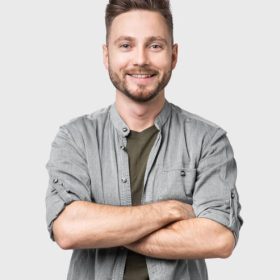 Cheerful young man with crossed hands looking to the camera. Isolated on gray background